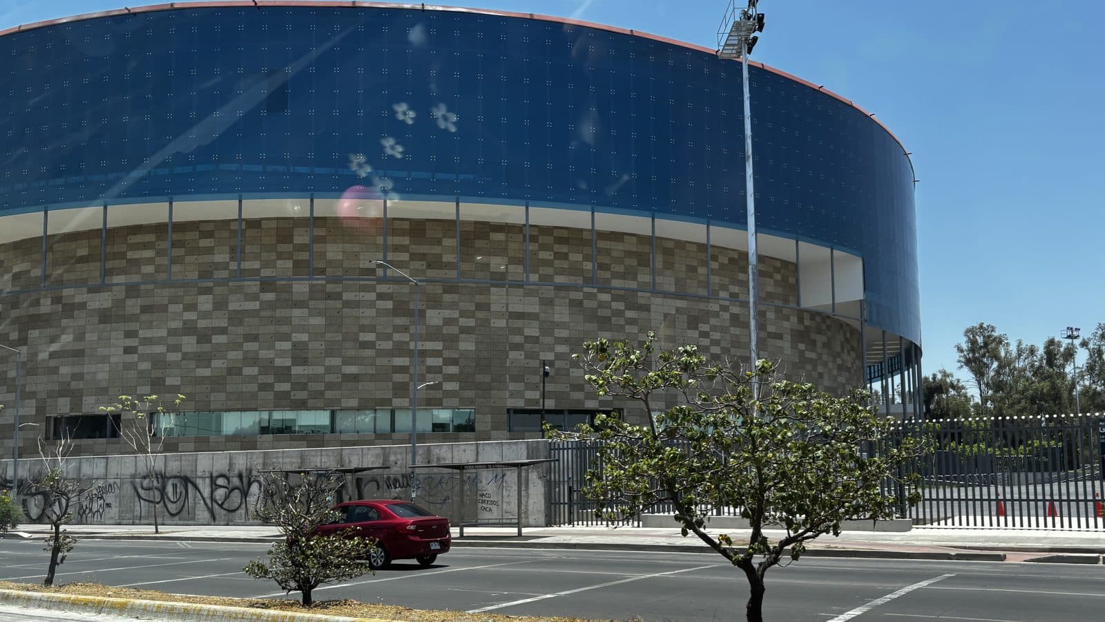 Exterior de la Arena Guadalajara: un gran cilindro de vidrio azul sobre una base de piedra, visto desde la calle.