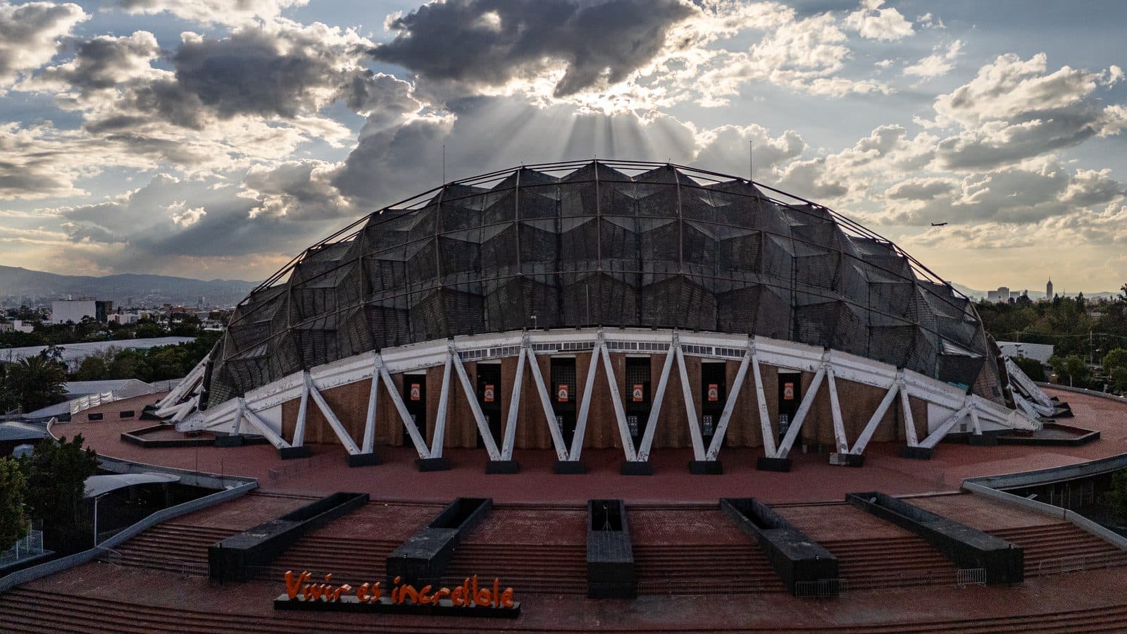Vista aérea del Palacio de los Deportes: la cúpula de cobre sobre sus arcos blancos, con nubes al atardecer.